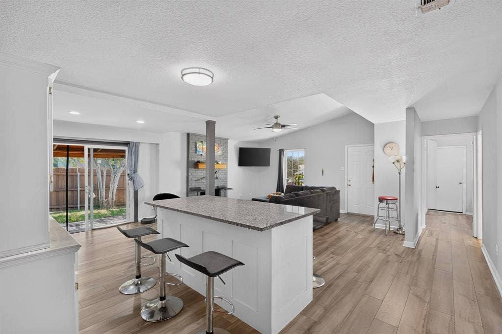 2228 Bunker Hill Drive, Carrollton, TX 75006 Open concept interior featuring wood-finish flooring, a central kitchen island with a stone-finish countertop, vaulted ceilings, and a fireplace with brick masonry