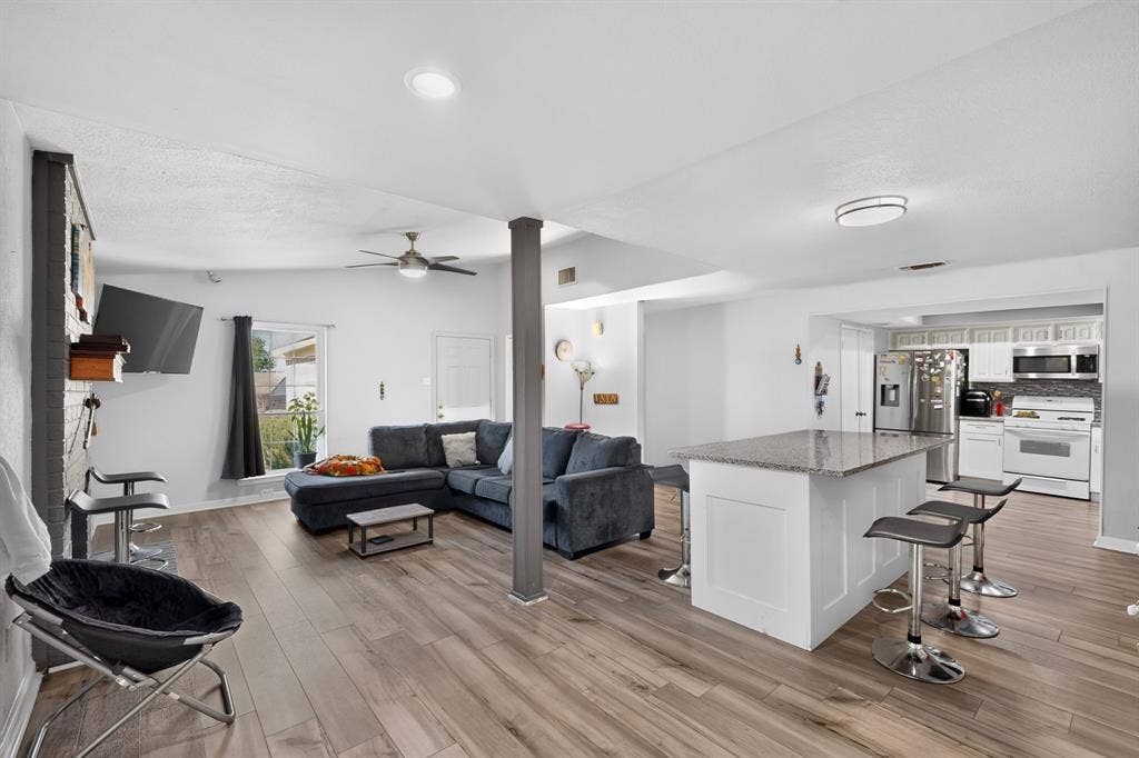 2228 Bunker Hill Drive, Carrollton, TX 75006 Open-concept interior featuring wood-finish flooring, a kitchen island with stone-finish countertop, white cabinetry, a structural support column, and a brick-faced fireplace surround