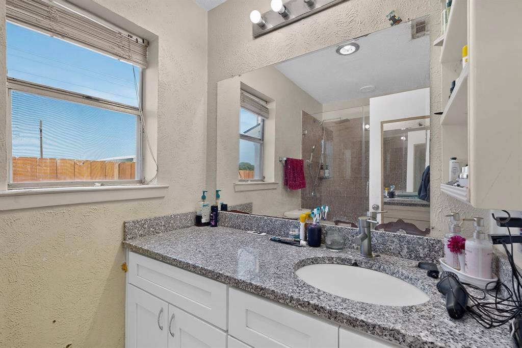 2228 Bunker Hill Drive, Carrollton, TX 75006 Bathroom featuring white cabinetry, stone-surface countertop with an integrated undermount sink, large wall-mounted mirror, and glass-enclosed shower