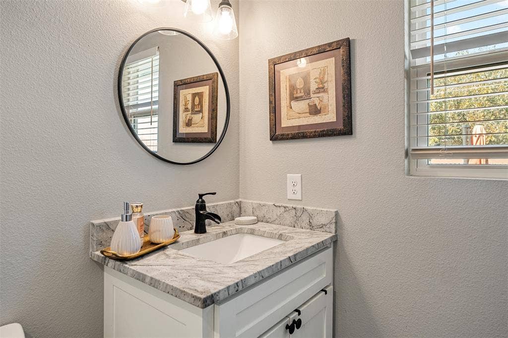 519 MAE Drive, Mabank, TX 75147 Vanity featuring a white cabinet, patterned stone countertop with an integrated rectangular sink, matte black faucet, and a circular wall-mounted mirror