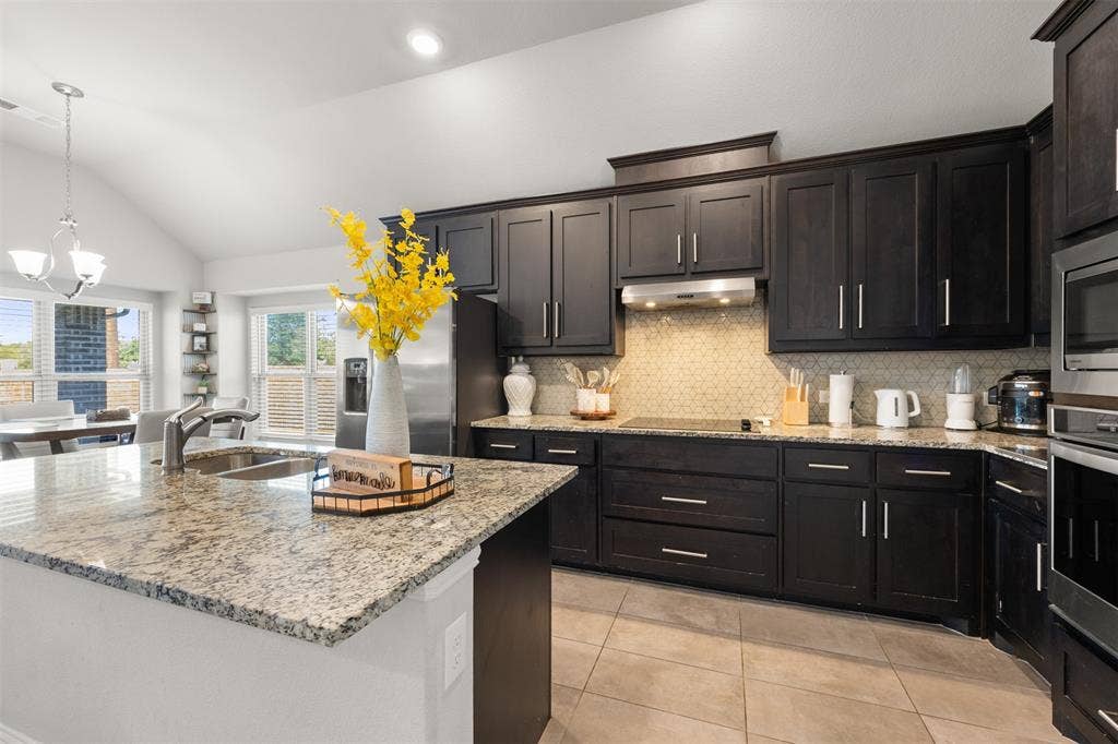 Kitchen featuring dark-finish cabinetry, stone-surface countertops, neutral tile backsplash, stainless steel built-in appliances, and vaulted ceilings