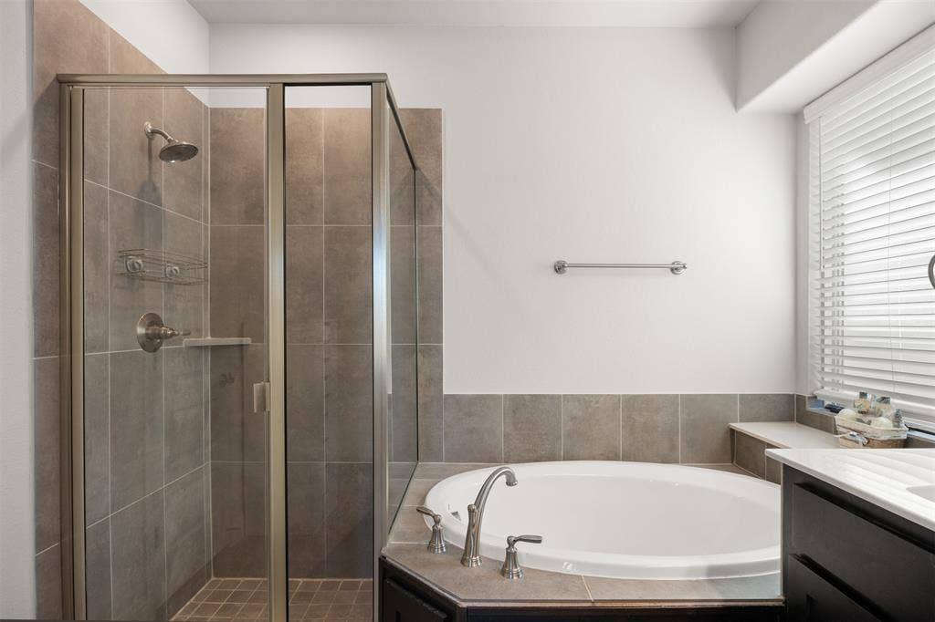 Corner soaking tub with tiled surround, walk-in shower with glass enclosure, large-format wall tile, polished metal fixtures, and integrated vanity countertop