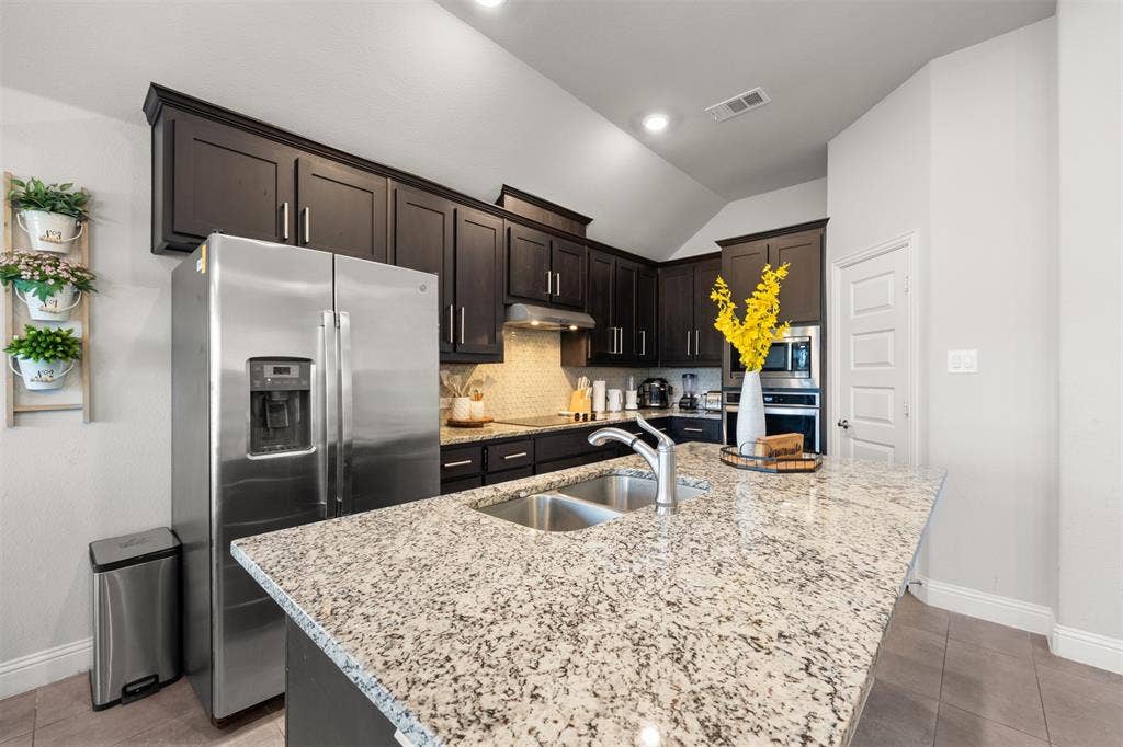 Kitchen featuring dark-stained cabinetry, stone-finish countertops, stainless steel appliances, a dual-basin sink with a gooseneck faucet, and vaulted ceilings