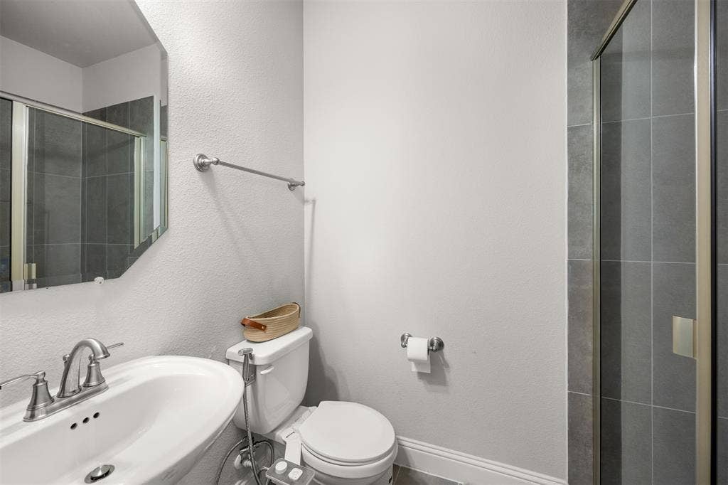 Bathroom featuring a white pedestal sink with a metallic faucet, a geometric wall mirror, and a glass-enclosed shower with large-format grey tile surround