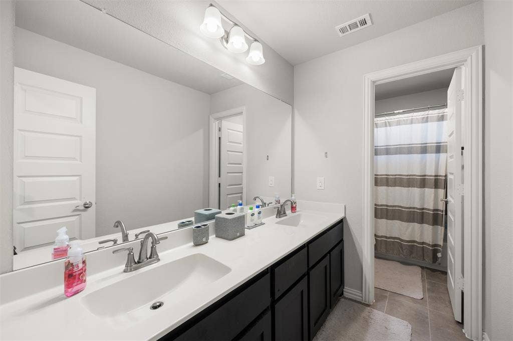 Double vanity with white solid-surface countertops, dark cabinetry, full-width wall mirror, and three-bulb vanity light fixture