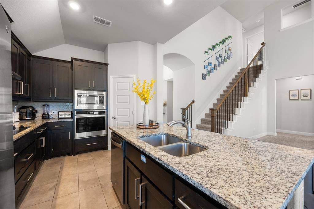 Kitchen featuring dark-finish cabinetry, stone-style countertops, stainless steel double-oven, and a central island with a dual-basin sink