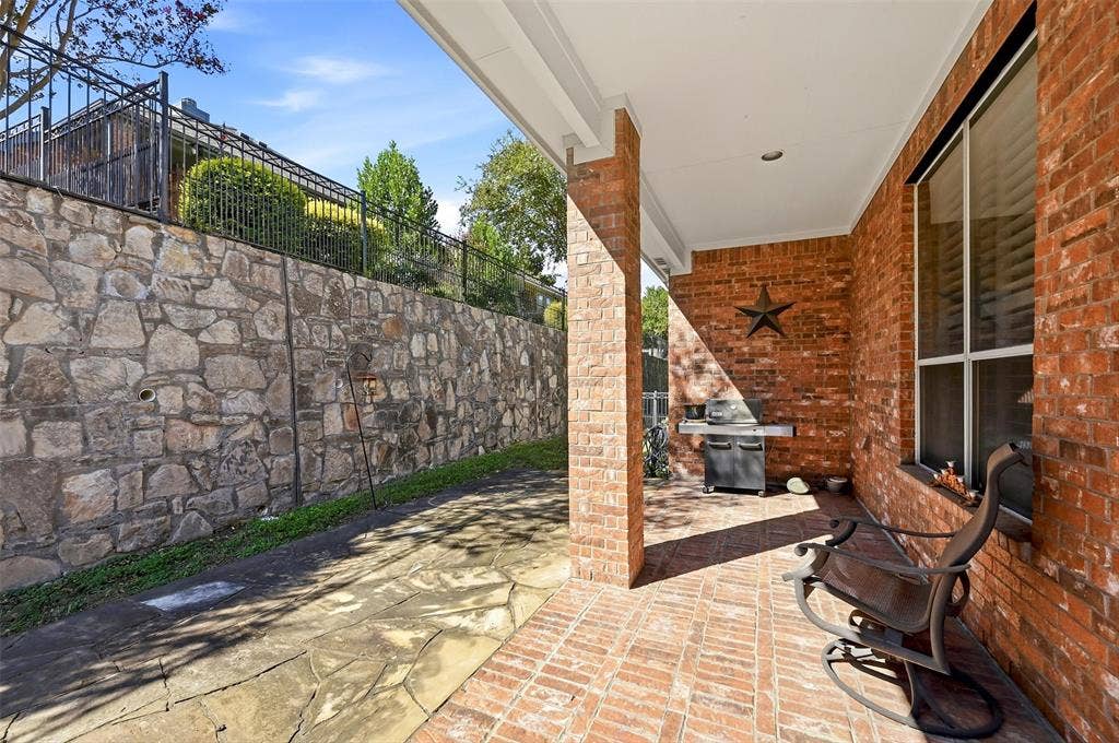 819 Mustang Drive, Fairview, TX 75069 Covered patio featuring red brick flooring and a masonry column, complemented by a natural stone retaining wall and a black metal perimeter fence