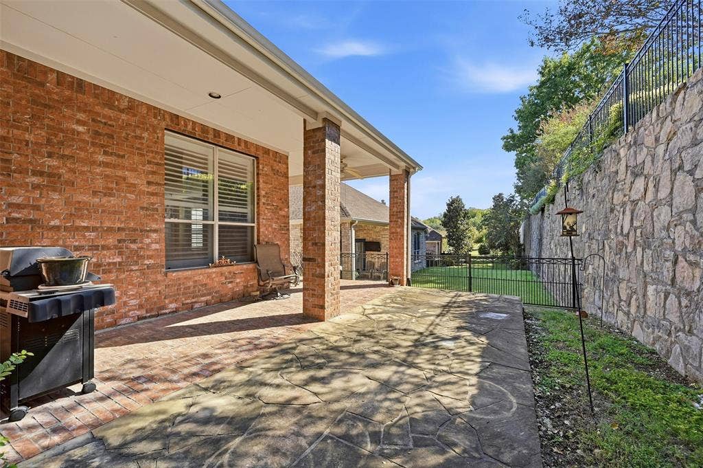 819 Mustang Drive, Fairview, TX 75069 Covered patio with brick exterior and stone paver flooring, featuring a large tiered stone retaining wall and metal perimeter fencing