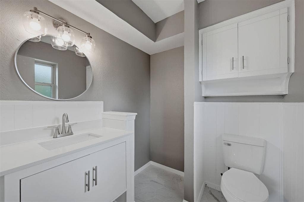 607 N Louisiana Drive, Celina, TX 75009 White vanity with rectangular undermount sink, oval wall-mounted mirror, three-bulb vanity light fixture, white wall-mounted storage cabinetry, and half-wall wainscoting