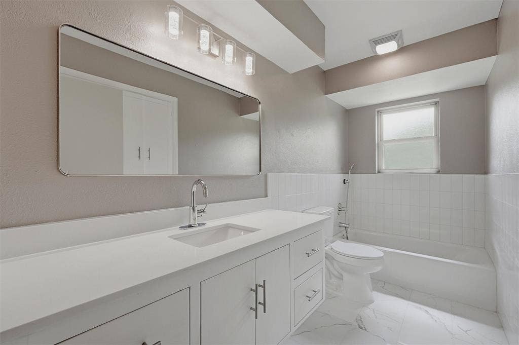 607 N Louisiana Drive, Celina, TX 75009 Bathroom featuring a white vanity with integrated rectangular sink and stone-finish countertop, large wall-mounted rectangular mirror, four-light vanity fixture, white tile tub surround, and tile flooring