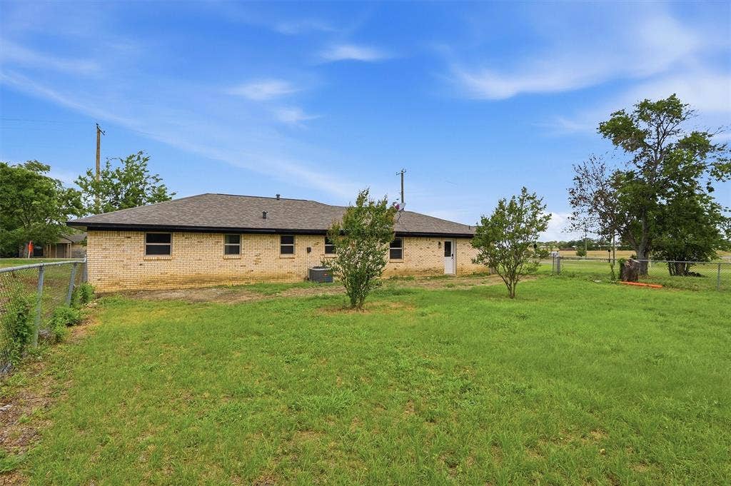 607 N Louisiana Drive, Celina, TX 75009 Exterior view featuring a tan brick facade, hip roof with composition shingles, metal chain-link perimeter fencing, and mature trees on a level grass lot