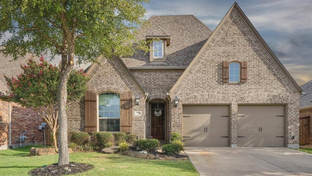 124 Village Creek Drive, McKinney, TX 75071 Brick exterior featuring a high-pitched roofline, arched entryway, decorative wood shutters, and an attached two-car garage