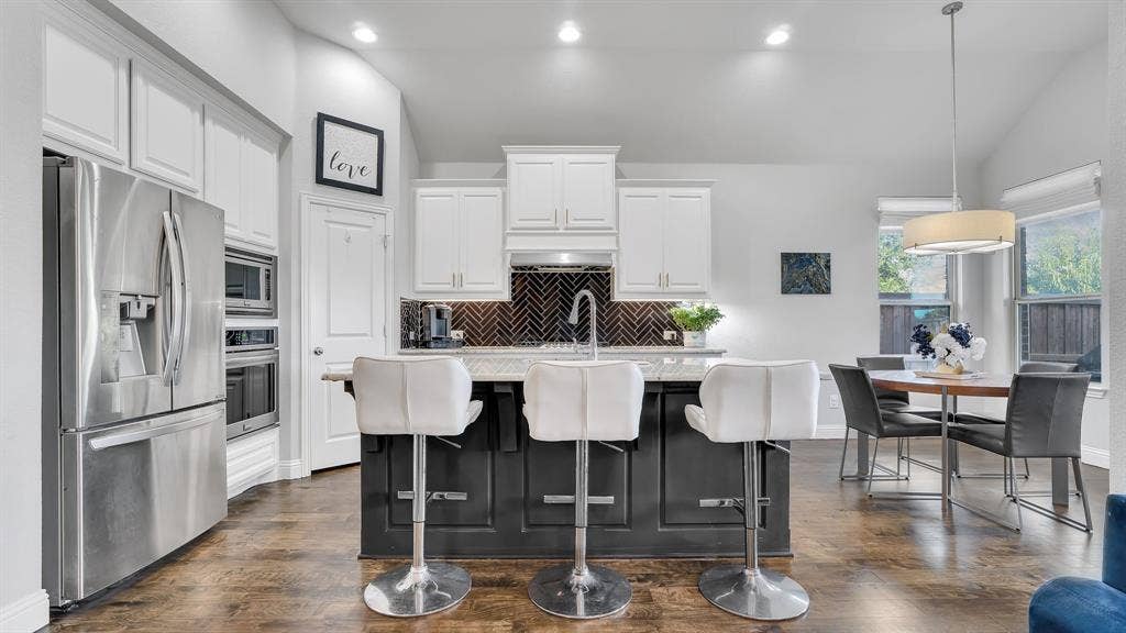 124 Village Creek Drive, McKinney, TX 75071 Kitchen featuring a central island with a contrasting dark base, white cabinetry, stainless steel appliances, herringbone-patterned tile backsplash, and wood-finish flooring