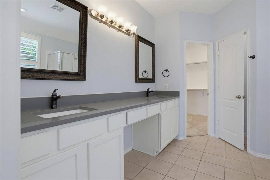 1402 Bradford Trace Drive, Allen, TX 75002 Double-vanity cabinet with white shaker-style doors, matte gray solid-surface countertop, dual under-mount sinks with dark metal faucets, and multi-light vanity fixtures