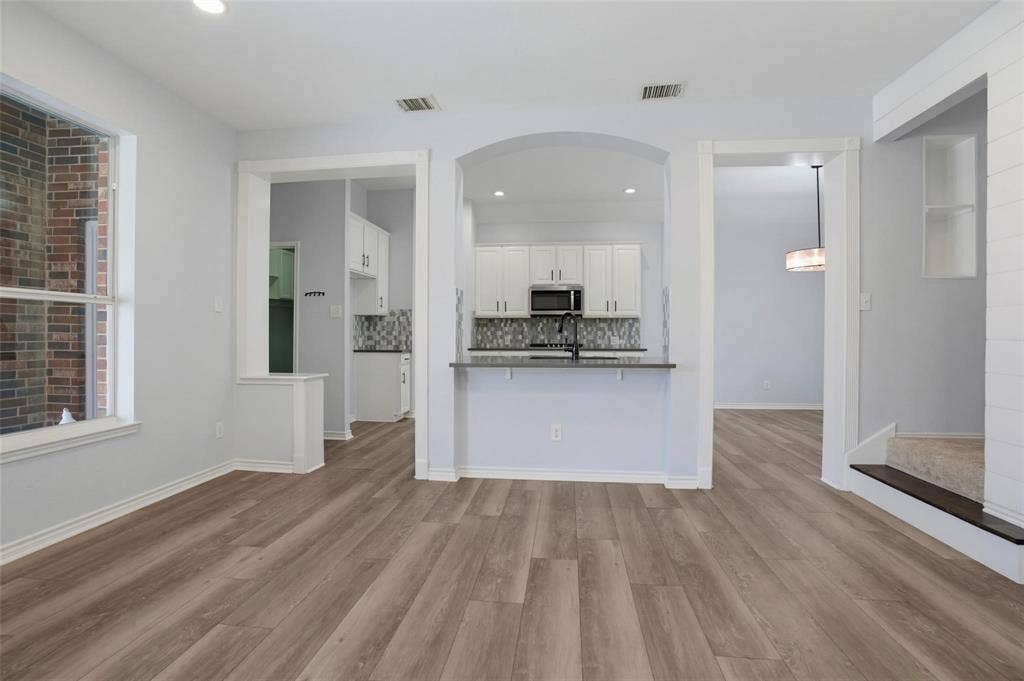 1402 Bradford Trace Drive, Allen, TX 75002 Interior space featuring wood-finish flooring, an arched kitchen entryway, white cabinetry, a breakfast bar with stone-finish countertops, and an exposed brick accent wall