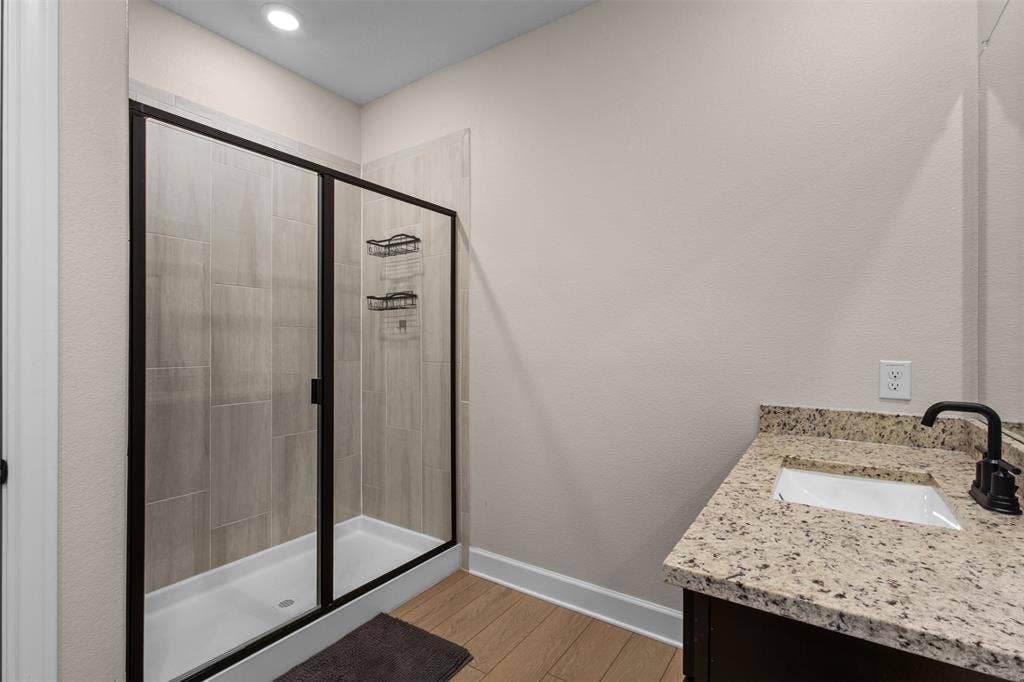 Walk-in shower with floor-to-ceiling tile surround and black metal frame, stone-finish countertop, undermount sink, matte black faucet, and wood-finish flooring