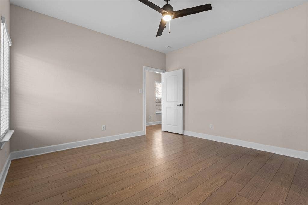 Neutral-toned room featuring wood-finish flooring, a ceiling fan with integrated lighting, and a two-panel interior door with matte black hardware