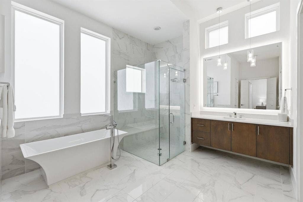 7853 Element Avenue, Plano, TX 75024 Freestanding soaking tub, glass-enclosed shower with bench seating, dual-sink wood vanity, large backlit mirror, and floor-to-ceiling stone-style wall tile