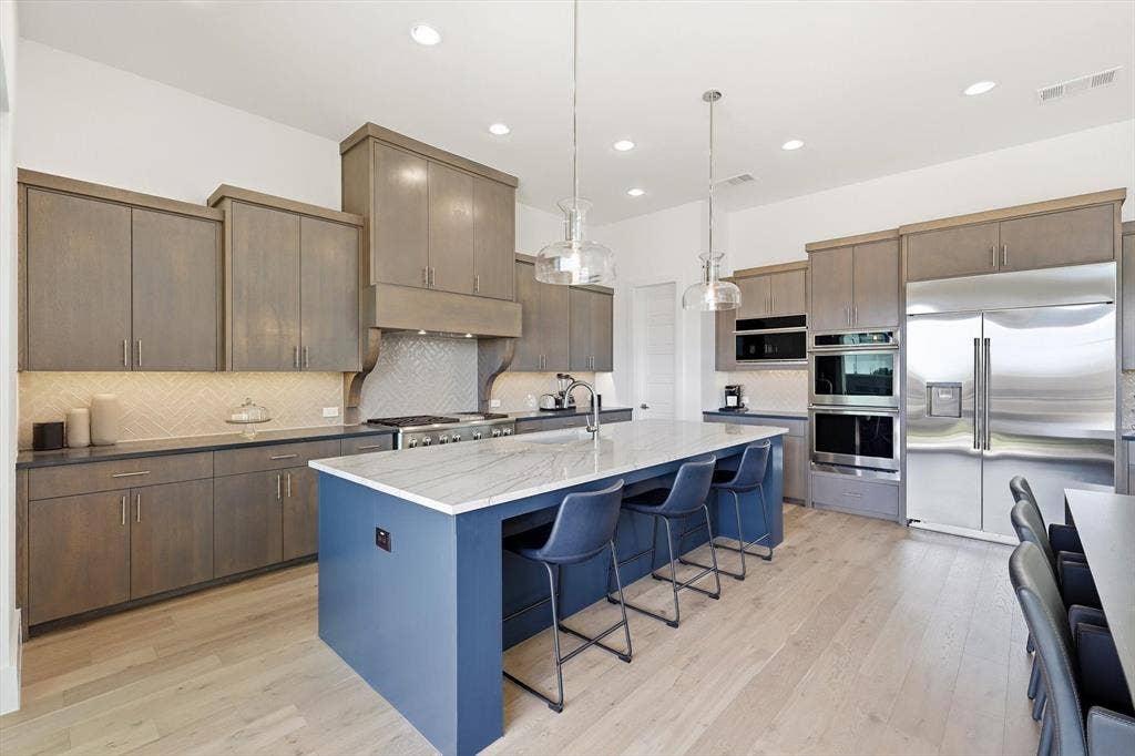 7853 Element Avenue, Plano, TX 75024 Kitchen featuring custom stained cabinetry, a central island with stone-finish countertops, herringbone tile backsplash, recessed lighting, and stainless steel built-in appliances