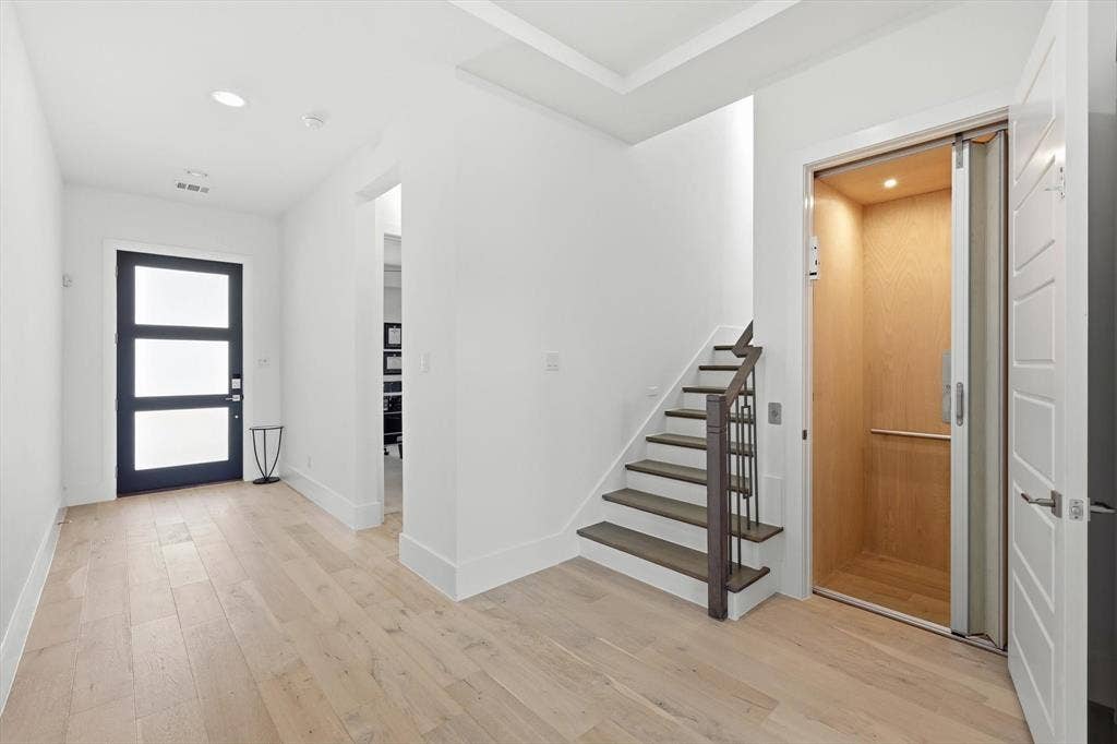 7853 Element Avenue, Plano, TX 75024 Entryway featuring light-toned wood-finish flooring, a dark-framed multi-pane glass door, recessed ceiling lighting, a staircase with dark wood treads and a coordinating metal railing, and a built-in elevator with wood-panel interior finishes