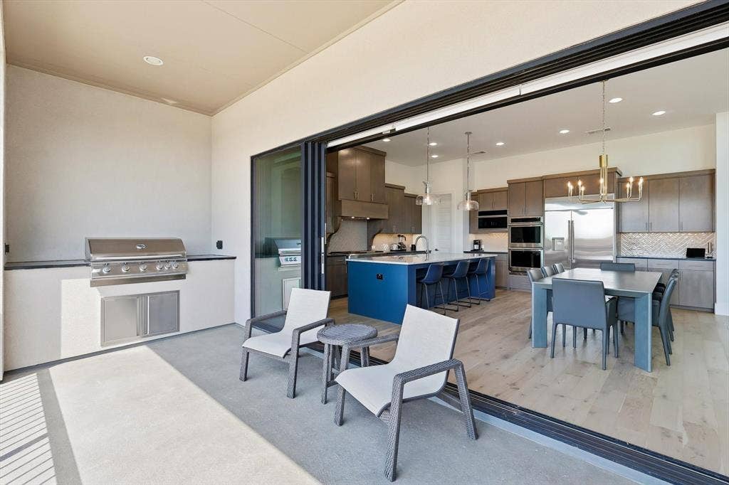 7853 Element Avenue, Plano, TX 75024 Covered patio featuring a built-in stainless steel grill, retractable glass wall system, wood-finish flooring, central kitchen island with seating, and dual wall-mounted ovens