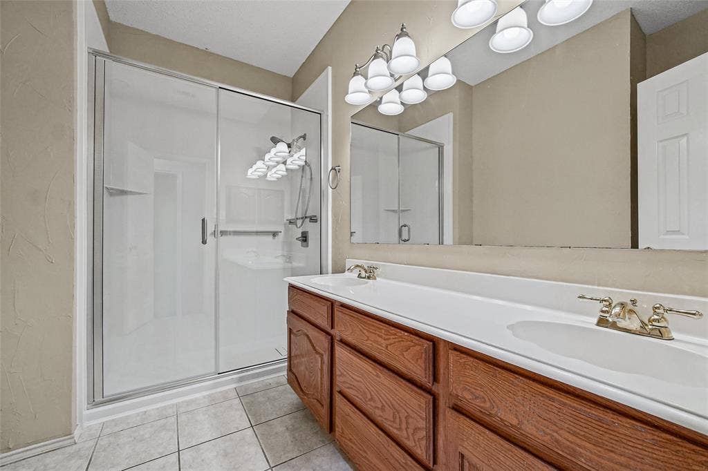 714 Cottonwood Bend Drive, Allen, TX 75002 Bathroom featuring a double-sink vanity with wood-finish cabinetry, a full-width wall mirror, a glass-enclosed shower with a sliding door, and neutral tile flooring