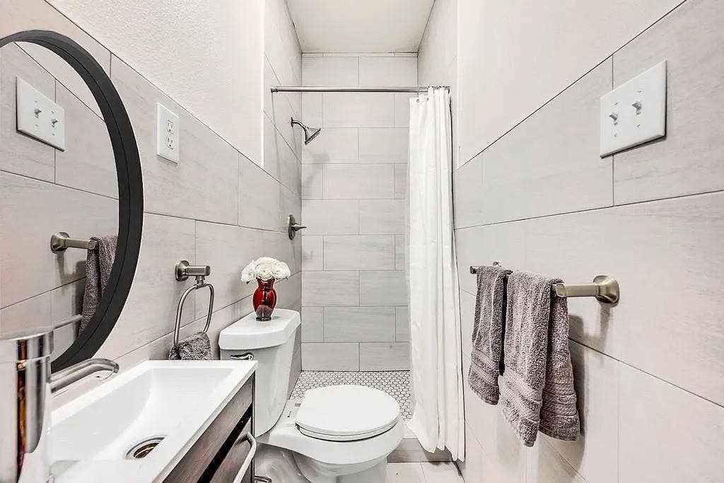 Full-wall horizontal tiling, oval vanity mirror with a black frame, wall-mounted faucet, shower with metallic hardware, and mosaic floor tiling