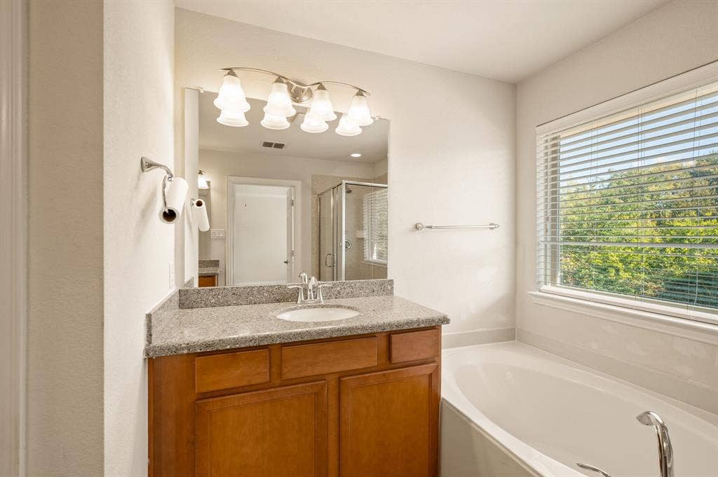 1712 Coronado Street, Allen, TX 75013 Bathroom vanity featuring a stone-composite countertop, undermount sink, multi-bulb light fixture, and deep-toned wood cabinetry, accompanied by a built-in soaking tub