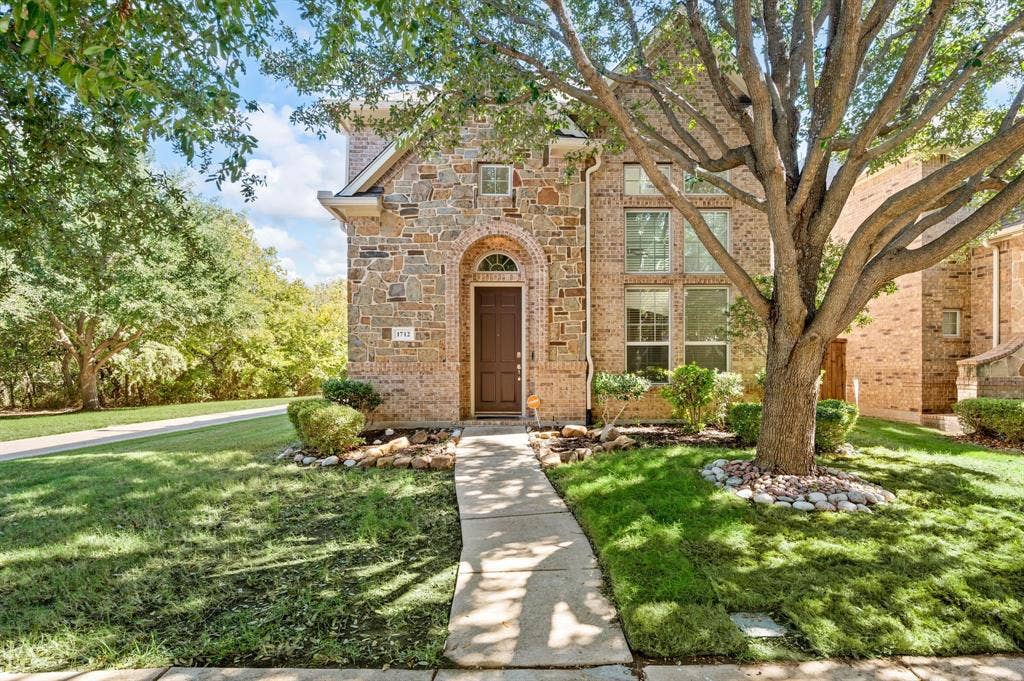 1712 Coronado Street, Allen, TX 75013 Multi-story stone masonry exterior featuring an arched entryway, tall multi-pane windows, and a mature tree-lined front yard with stone-bordered landscaping