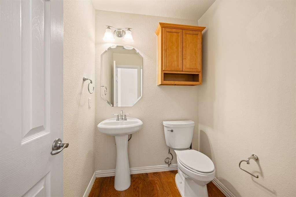 1712 Coronado Street, Allen, TX 75013 Pedestal sink with polished chrome faucet, octagonal wall mirror, dual-light vanity fixture, wooden wall-mounted storage cabinet, and wood-finish flooring