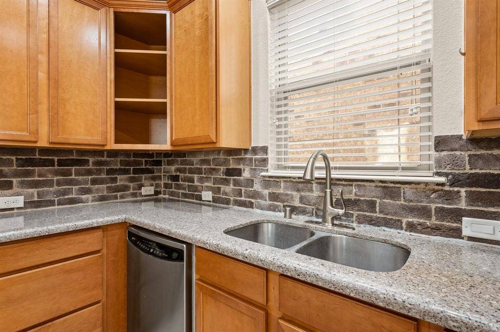 1712 Coronado Street, Allen, TX 75013 Kitchen area featuring stained wood cabinetry, stone-surface countertops, a dark-toned brick backsplash, a stainless steel double-basin undermount sink, and a high-arch pull-down faucet