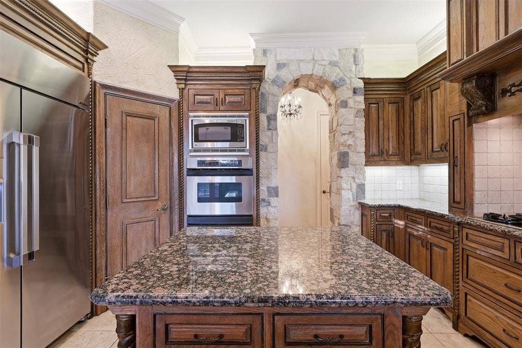 1919 Kings Pass, Heath, TX 75032 Gourmet kitchen featuring a granite-topped island, rich wood cabinetry, stainless steel appliances, a stone archway, and tile backsplash