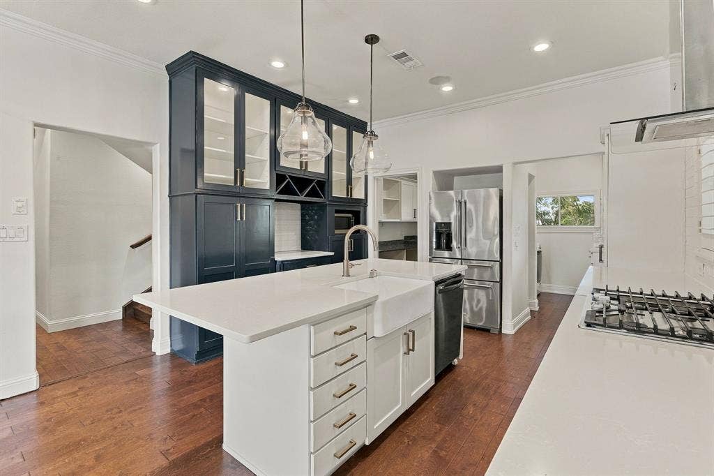 510 E Malone Street, Celina, TX 75009 Kitchen featuring a central island with a farmhouse-style sink, white solid-surface countertops, floor-to-ceiling cabinetry with glass-front uppers, stainless steel appliances, and wood-finish flooring