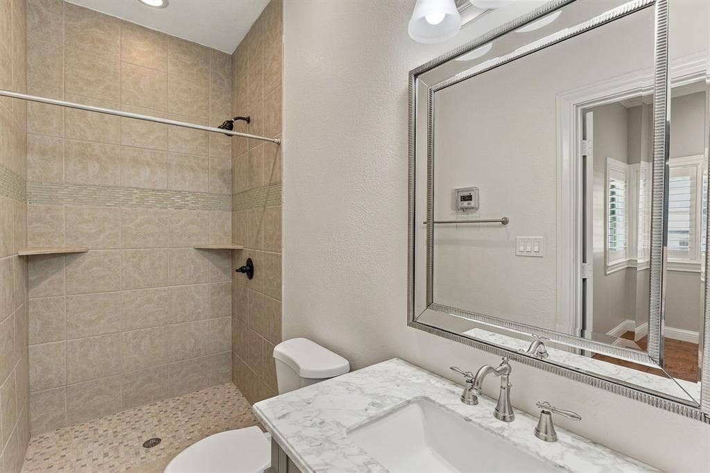 510 E Malone Street, Celina, TX 75009 Bathroom featuring a vanity with stone-finish countertop, undermount sink, and integrated mirror, accompanied by a tiled walk-in shower with decorative glass inlay and pebble-texture flooring