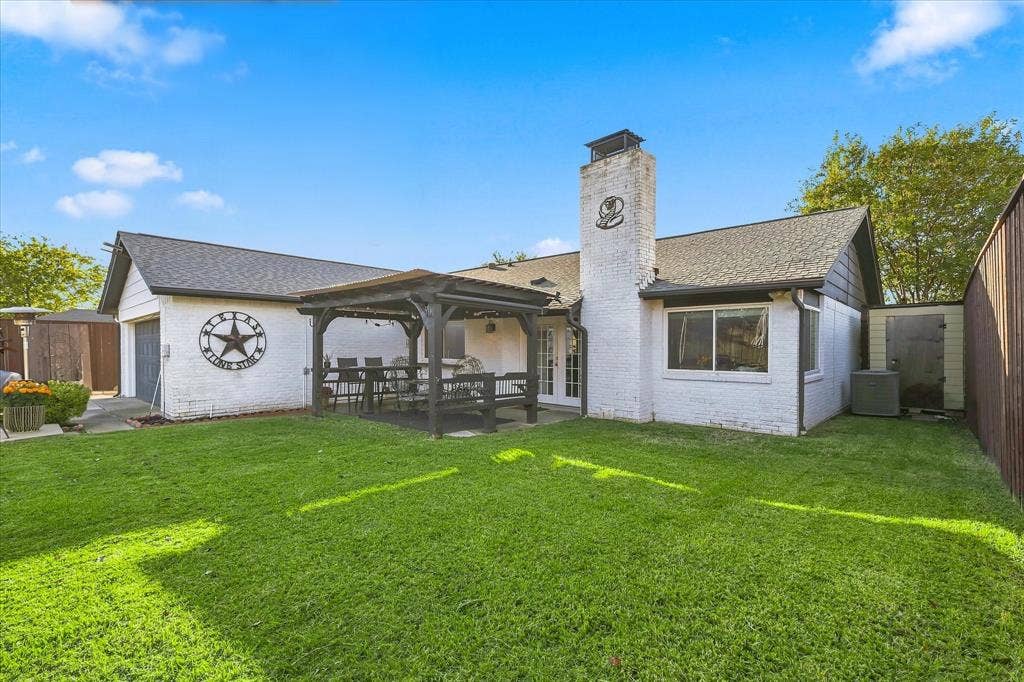 2117 Northmoor Drive, Carrollton, TX 75006 Fenced backyard featuring a manicured lawn, a covered timber pergola, a prominent masonry chimney, and white brick exterior finish