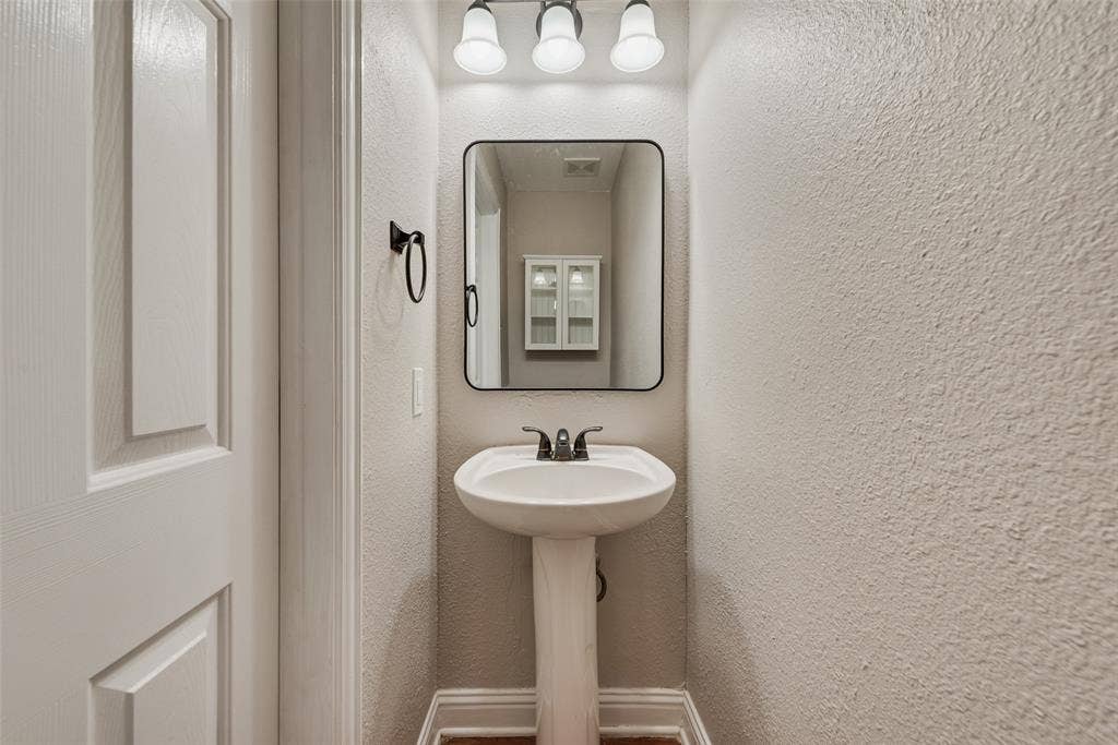 2017 Lunenburg Drive, Allen, TX 75013 Pedestal sink with a dark metal faucet, rectangular mirror with a black frame, and a three-light fixture with white glass shades