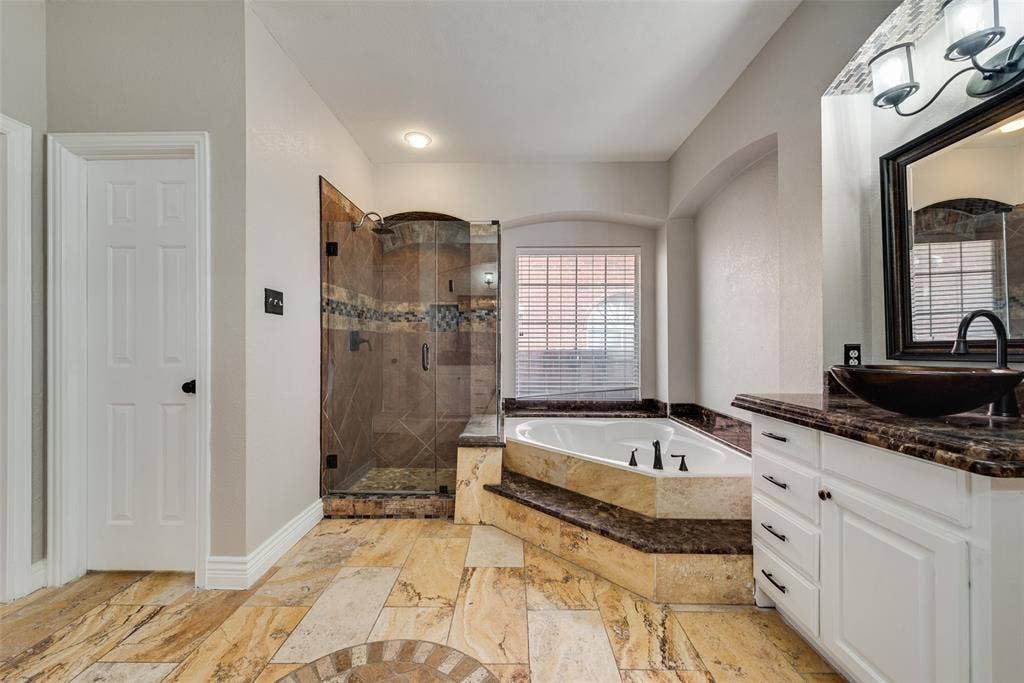 2017 Lunenburg Drive, Allen, TX 75013 Bathroom featuring stone-tile flooring, a jetted soaking tub with a tiled surround, a glass-enclosed shower with decorative inlay, and a vanity with a vessel sink