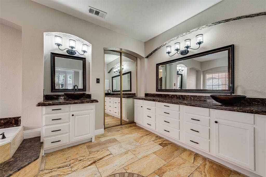 2017 Lunenburg Drive, Allen, TX 75013 Dual-vanity bathroom featuring white shaker-style cabinetry, dark stone countertops, vessel sinks, framed wall mirrors with integrated lighting, and natural stone flooring