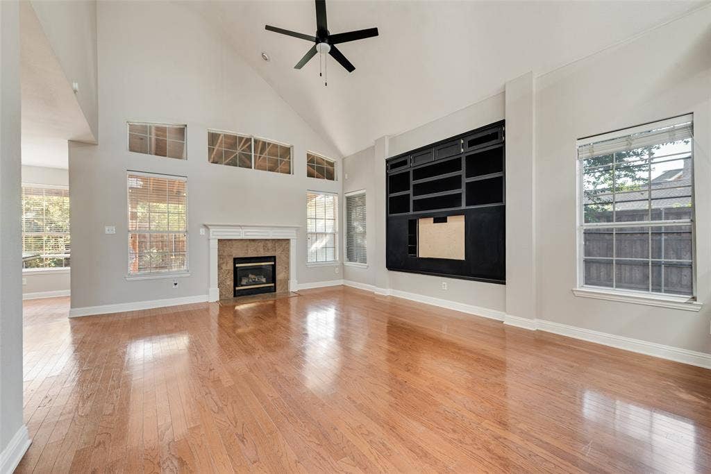 2017 Lunenburg Drive, Allen, TX 75013 Vaulted ceiling living area featuring wood-finish flooring, a fireplace with a stone-surround mantel, built-in black cabinetry, and oversized windows with transom accents