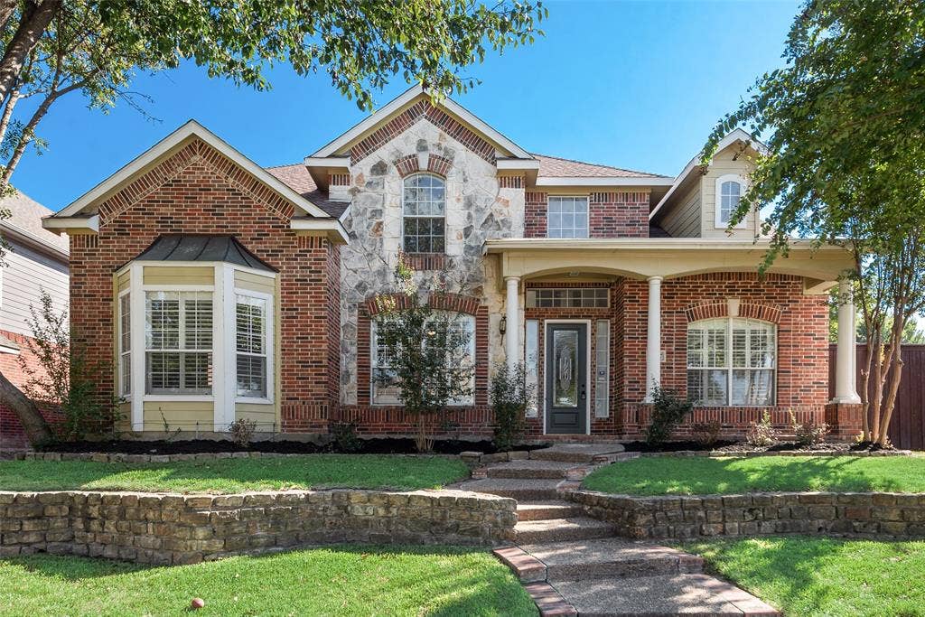 2017 Lunenburg Drive, Allen, TX 75013 Traditional brick exterior featuring stone veneer accents, a covered front porch with classical columns, a multi-paneled glass front door, and a tiered natural stone retaining wall