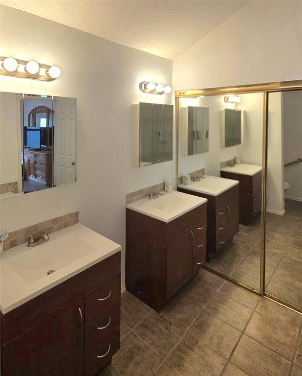 9436 Penny Lane, Irving, TX 75063 Dual dark-finish vanities with rectangular integrated sinks, stone-finish countertops, tiled backsplashes, and large mirrored closet doors with gold-tone frames over stone-tile flooring