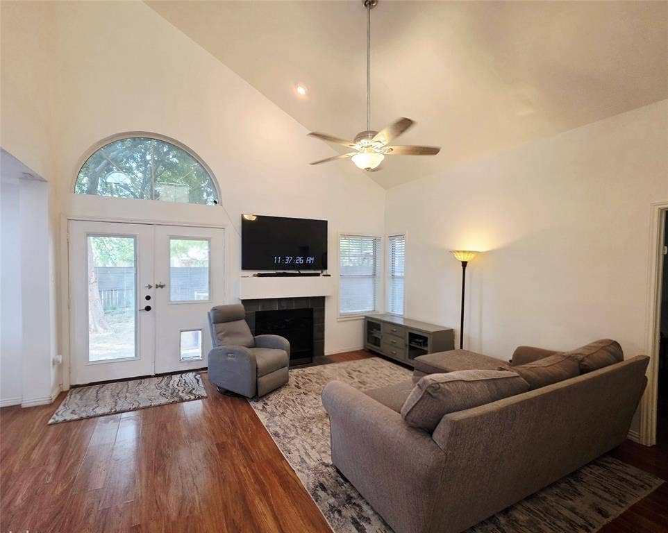 9436 Penny Lane, Irving, TX 75063 Vaulted ceiling with a central brushed metal ceiling fan, wood-finish flooring, fireplace with a tiled surround and mantel, and a multi-pane arch-top window above a double-entry door