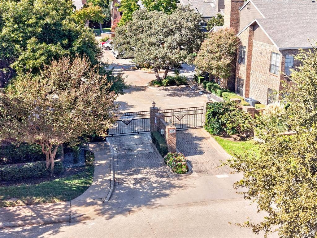 5859 Frankford Road, #1209, Dallas, TX 75252 Gated entry featuring a brick-pillared wrought iron gate, paved driveway, mature trees, and landscaping