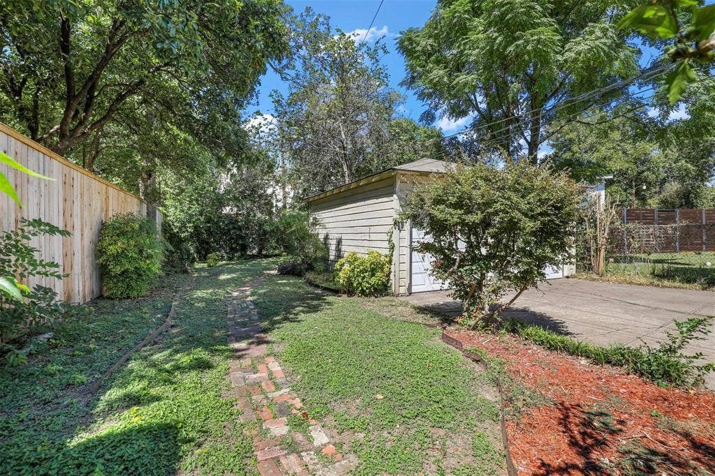 5925 Victor Street, Dallas, TX 75214 Rear exterior featuring a detached garage, concrete driveway, brick pathway, wood privacy fencing, and mature trees