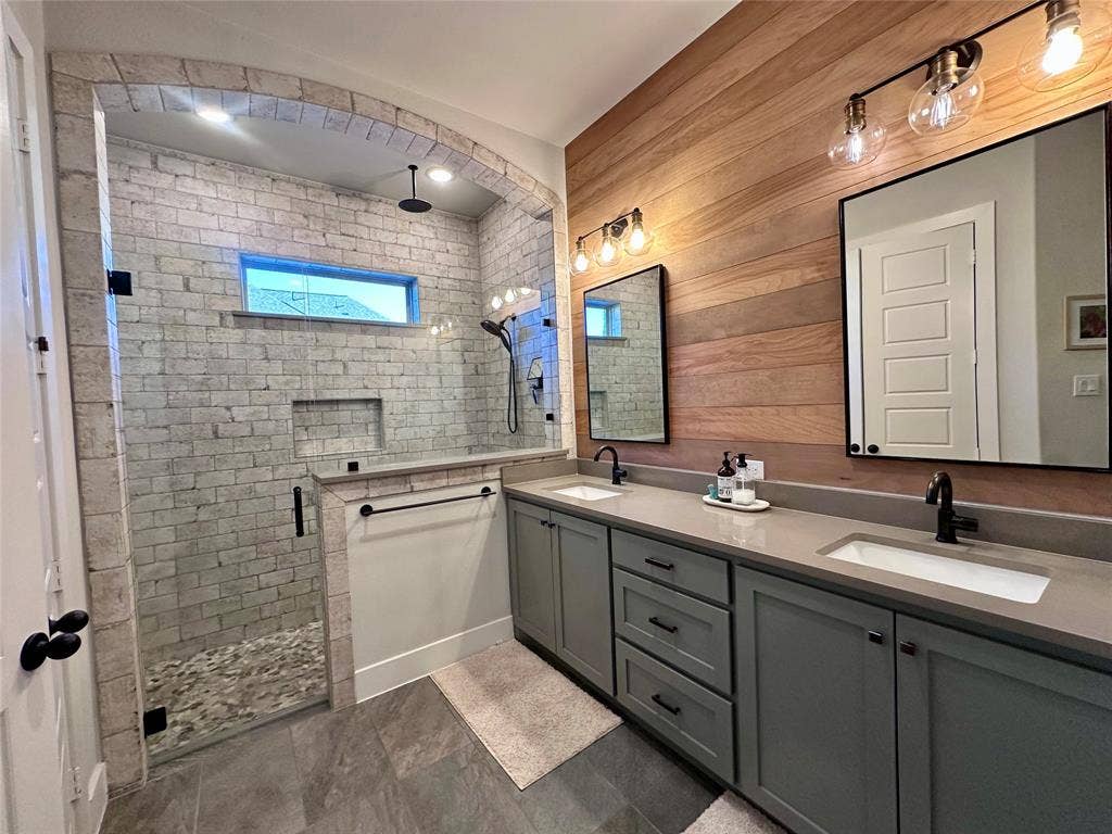 2712 War Admiral Street, Celina, TX 75009 Walk-in shower with brick-tiled walls and arched entry, double vanity with dual undermount sinks, wood-plank wall accent, and matte black hardware