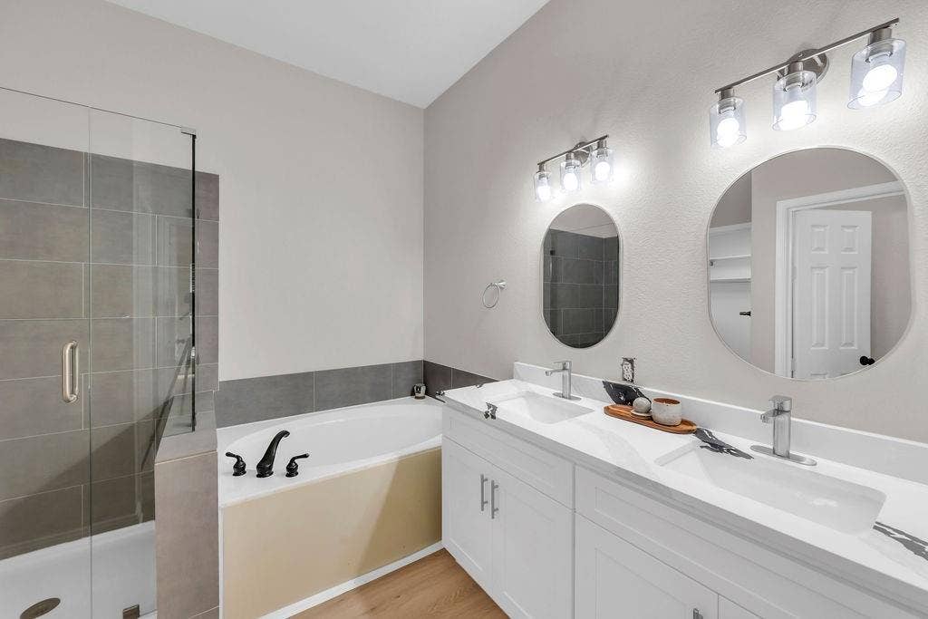 12109 Biloxi Drive, Frisco, TX 75035 Dual-sink vanity with white cabinetry and solid-surface countertop, walk-in shower with glass enclosure, deep soaking tub, and round mirrors with integrated three-light fixtures