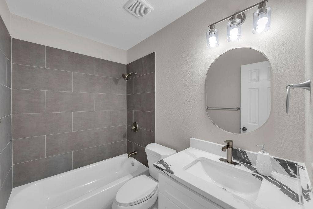 12109 Biloxi Drive, Frisco, TX 75035 Bathroom featuring a vanity with a patterned countertop, oval wall-mounted mirror, three-light fixture, and a tub-shower combination with floor-to-ceiling gray tile surround
