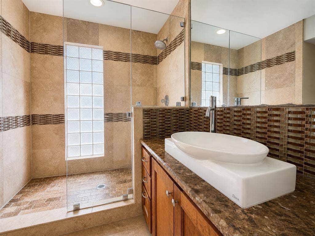 3210 Cortez Court W, Irving, TX 75062 Walk-in shower with a glass enclosure, glass block window, stone-finish tile surround, oval vessel sink, and stone-surface vanity countertop