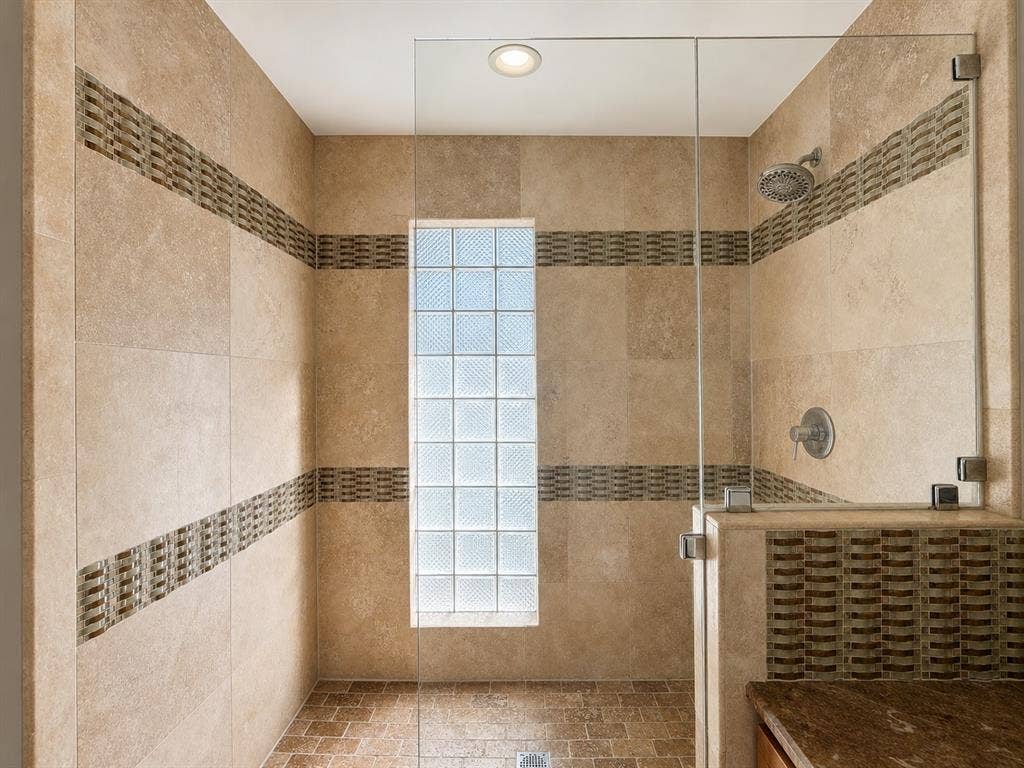 3210 Cortez Court W, Irving, TX 75062 Walk-in shower featuring natural stone wall tile, decorative mosaic tile accents, a glass block privacy window, frameless glass enclosure, and a wall-mounted showerhead