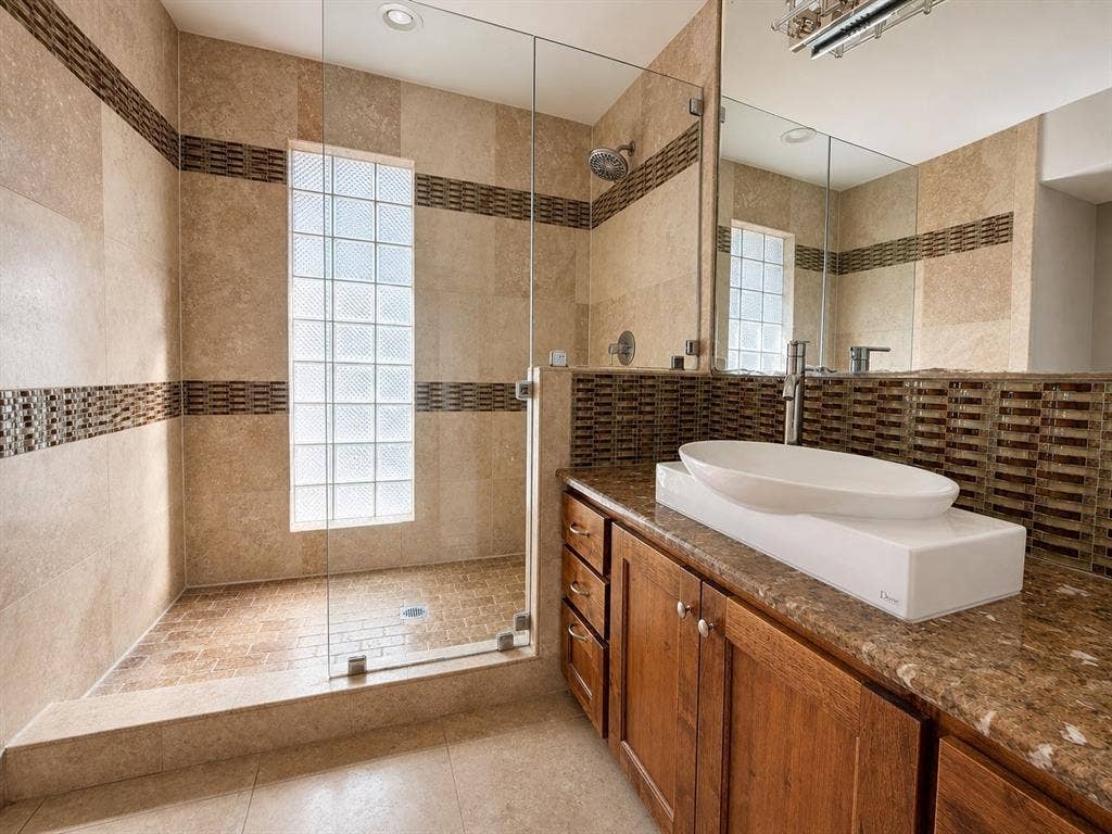 3210 Cortez Court W, Irving, TX 75062 Walk-in shower featuring glass paneling, glass block window, and horizontal accent tile, paired with a wood vanity, stone-finish countertop, and vessel sink